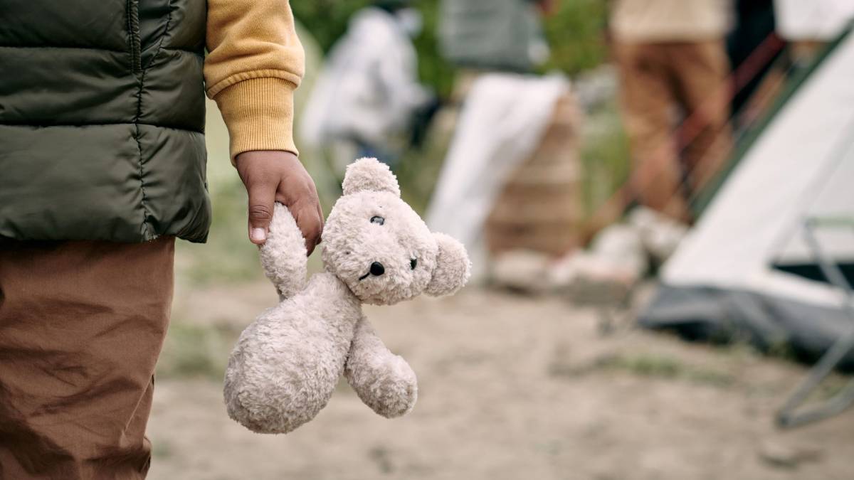 Ein Kind hält einen weißen Teddybären in seiner Hand. Im Hintergrund ist ein Zelt und Menschen zu sehen.