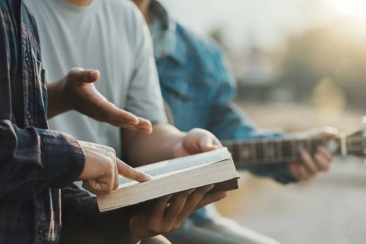 Mehrere Hände zeigen auf eine Bibel und gestikulieren. Im Hintergrund spielt jemand Gitarre.